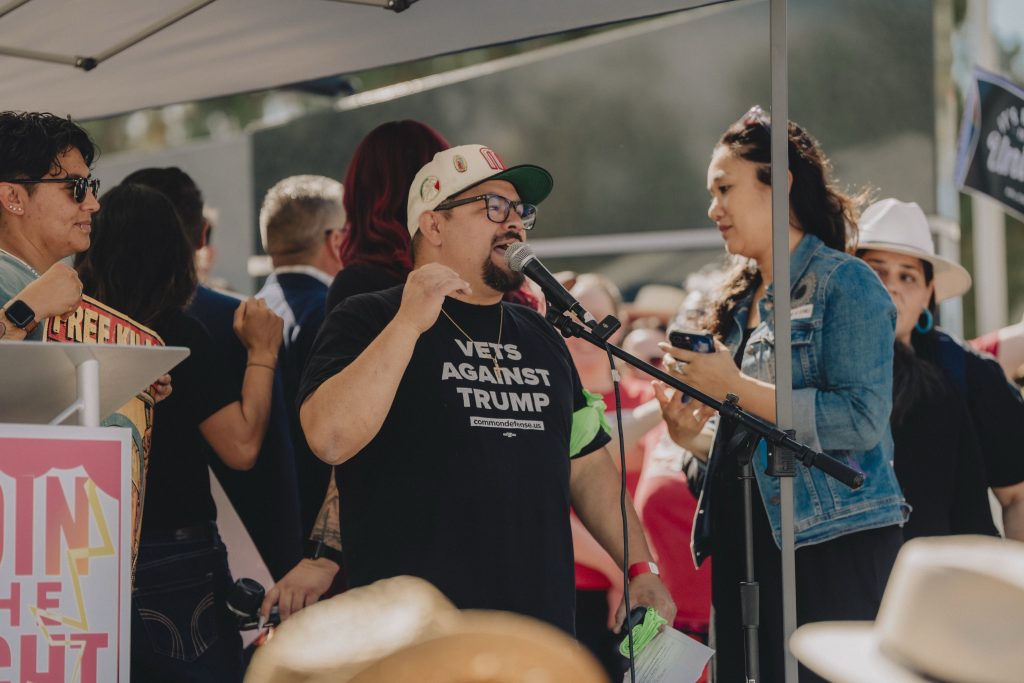 A man wearing a "Vets Against Trump" shirt speaks into a microphone at an outdoor gathering, surrounded by people.