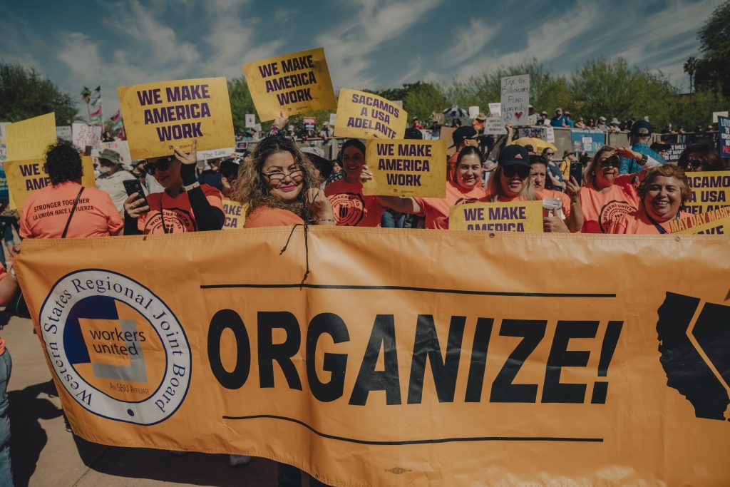 Protest gathering at Arizona State Capitol with focus on a group called "Workers United". The group is wearing orange shirts holding yellow signs with a banner in front with "ORGANIZE" in bold lettering.