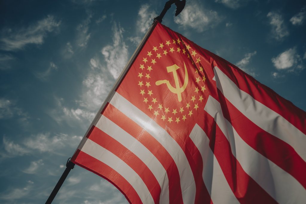 Image of U.S. flag up against blue slightly cloudy sky. Stars are replaced with the hammer and sickle logo surrounded with two rings of five-tipped stars in orange, against red.