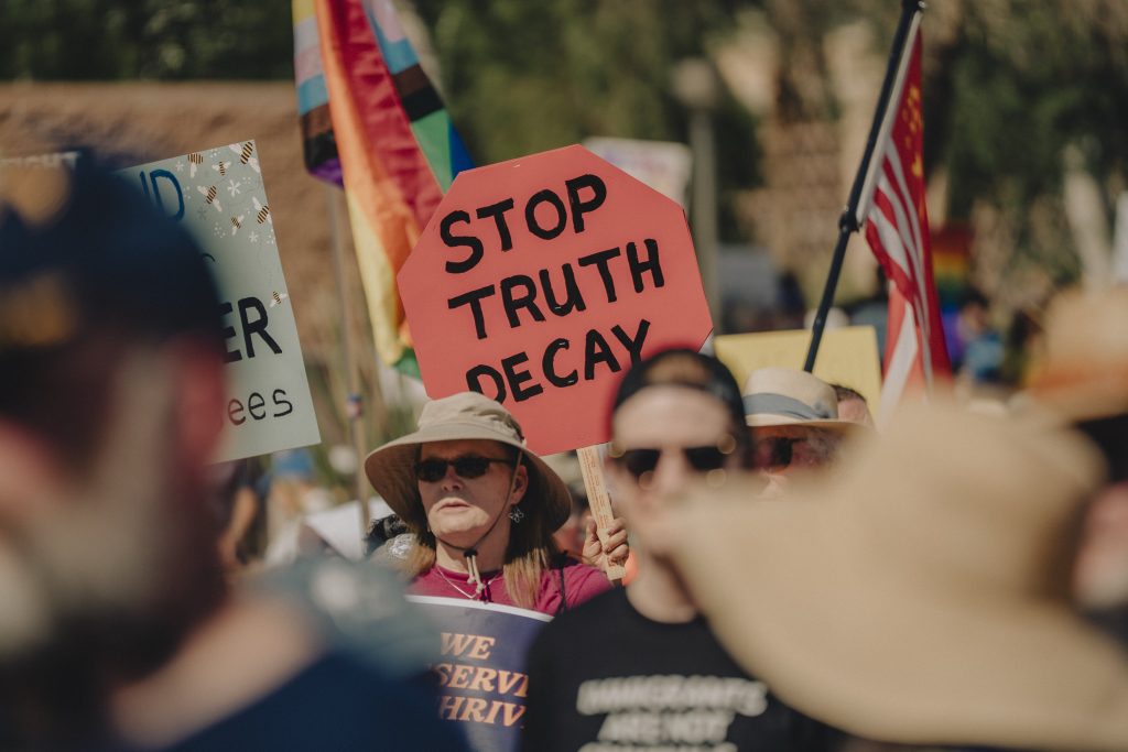 Zoomed-in image of protest gathering at Arizona State Capitol. One. LGBTQ+ flag and communist U.S. flag barely visible. One sign is readable. It is a red octagon that says "Stop truth decay"