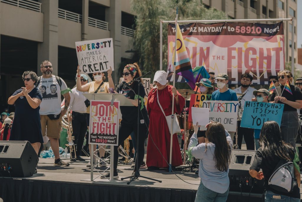 Picutre of woman speaking on stage with people holding signs behind her. She is reading from her phone. Sign language interpreter to her right. There are two monitors (speakers) on the stage. The woman is speaking at a podium with a sign that says "Join the fight. Text "AZ MAYDAY" to 58910 Now!"