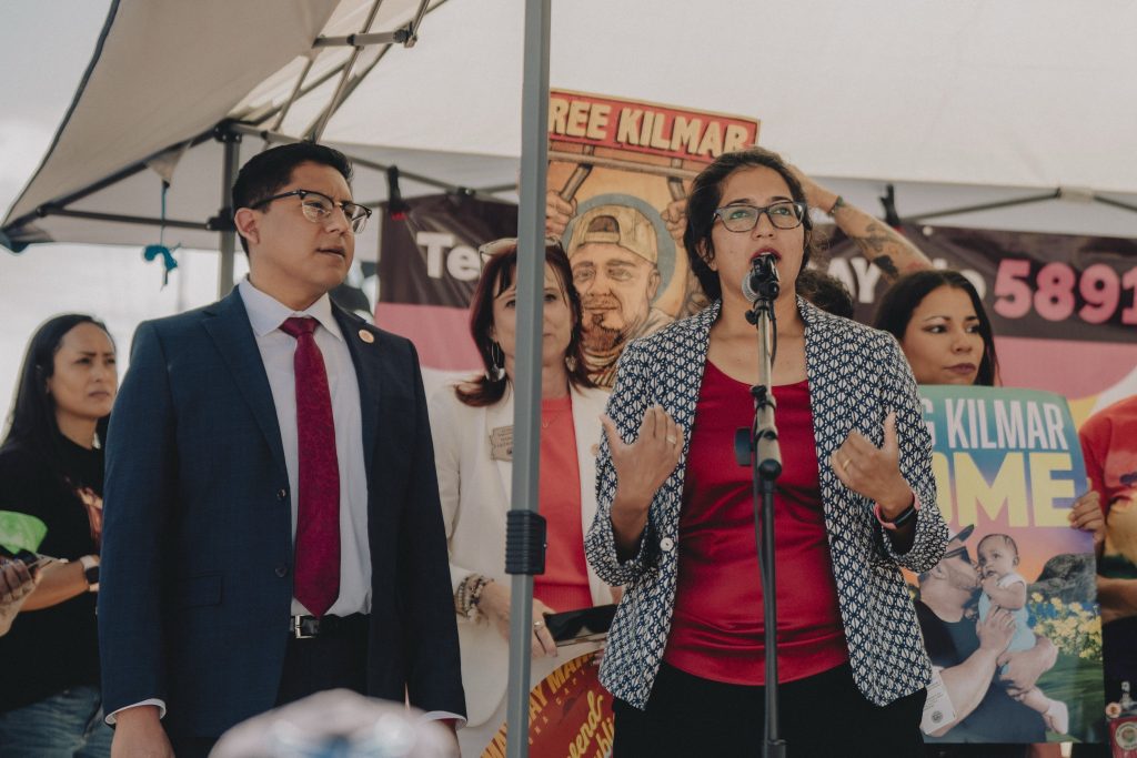 Woman speaking into microphone at a protest with people behind her and man to her right.