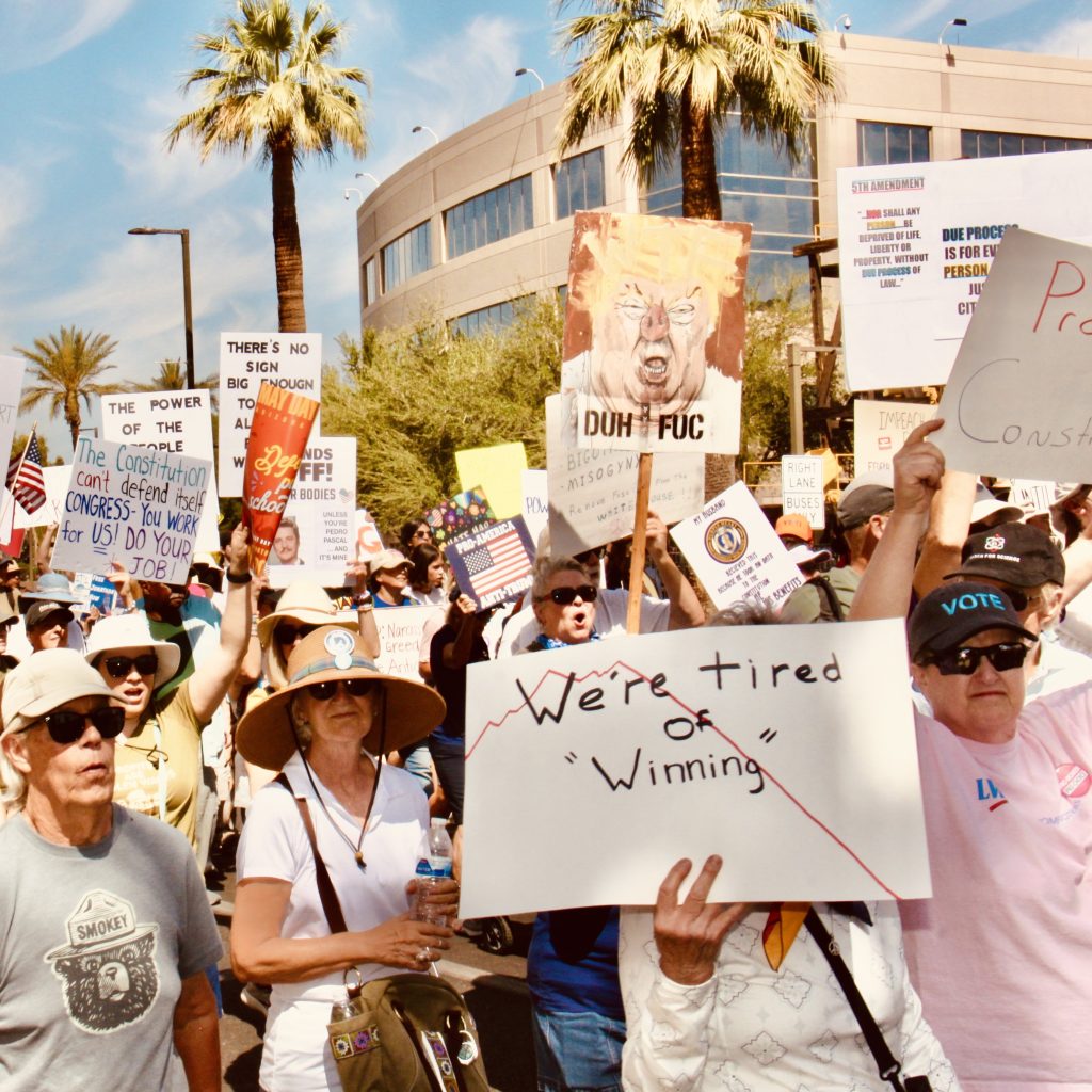 View of protest march with people holding up signs headed East from the Arizona State Capitol. Sings are as follows: "We're tired of winning" on top of a line chart going from jagged to angled downward; "DUH FUC" on top of an ugly painting of Donald Trump; "The Constitution can't defend itself. CONGRESS - You Work for US! DO YOUR JOB". In total about a dozen signs are visible along with a small U.S. flag.