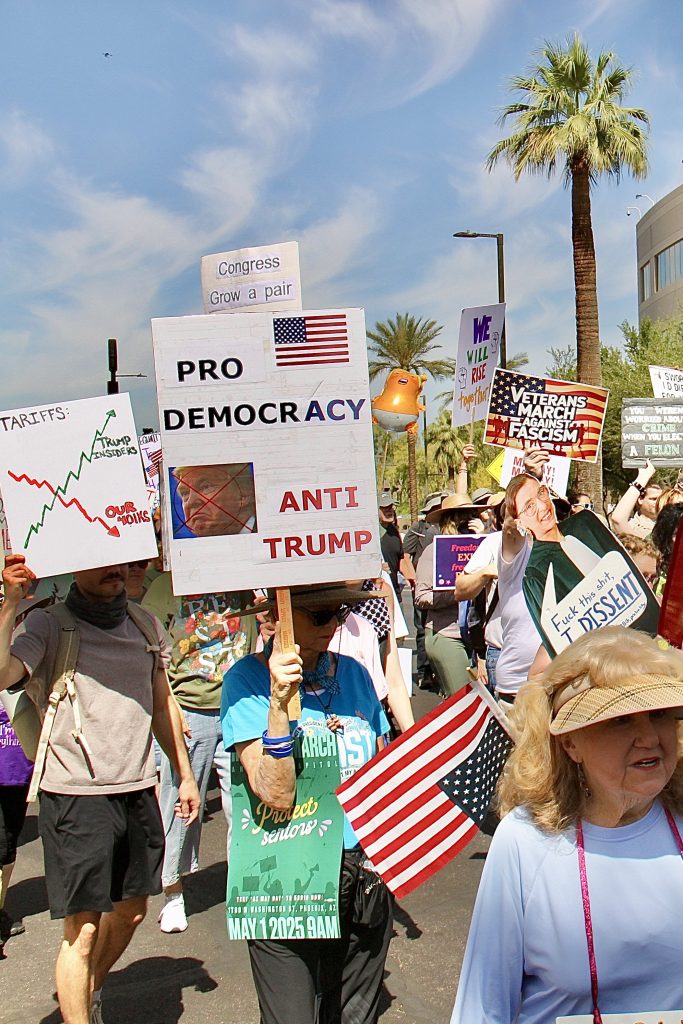 View of protest march with people holding up signs headed East from the Arizona State Capitol. Sings are as follows: U.S. Flag, "PRO DEMOCRACY", picture of trump crossed out "ANTI TRUMP" in red. "TARRIFFS:" green jagged line going upward labeled "Trump insiders", red jagged line labeled "Our bonus" "Veterans march against fascism" against a U.S. flag with soldiers silhouetted in between. "WE WILL RISE together" in different colors with two solidarity fists on the side" "Congress Grow a pair" cut off by first listed sign". U.S. flag on old lady's back Baby Trump balloon held up on hard stick About a dozen signs visible.