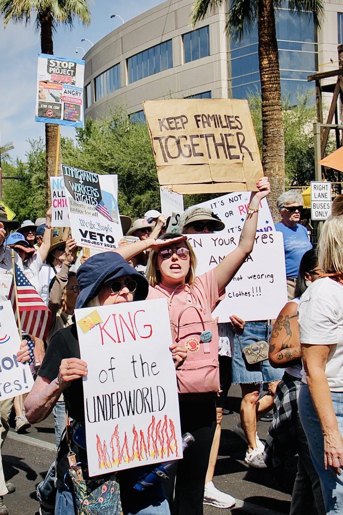 Protest march. About a dozen signs visible. Some of them are as follows: "KEEP FAMILIES TOGETHER" on tan paper "KING of the UNDERWORLD" with flames drawn on the bottom on white poster board"