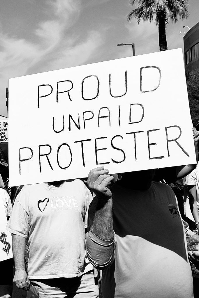 Black and white picture of protest march. One sign is visible: "PROUD UNPAID PROTESTER"