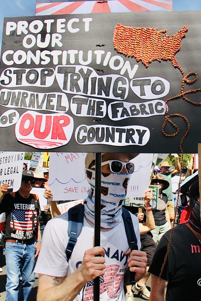Protest march focusing on man with white Old Navy t-shirt, and a Powerade cloth wrapped around his lower face and neck. Wearing a backpack and white sunglasses. Sign says: "Protect our constitution" "Stop trying to unravel the fabric of OUR country" with a mainland U.S. map made of multicolored yarn, with a strand coming off Florida, making loops, and hanging off the sign about 2 feet.