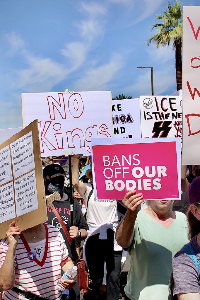 Protest march with about 5 signs visible, 2 fully readable.Frontmost one says "Bans off *our bodies* in white text on pink with some logo in bottom right. Other readable one says "NO Kings" in red outline text on white.