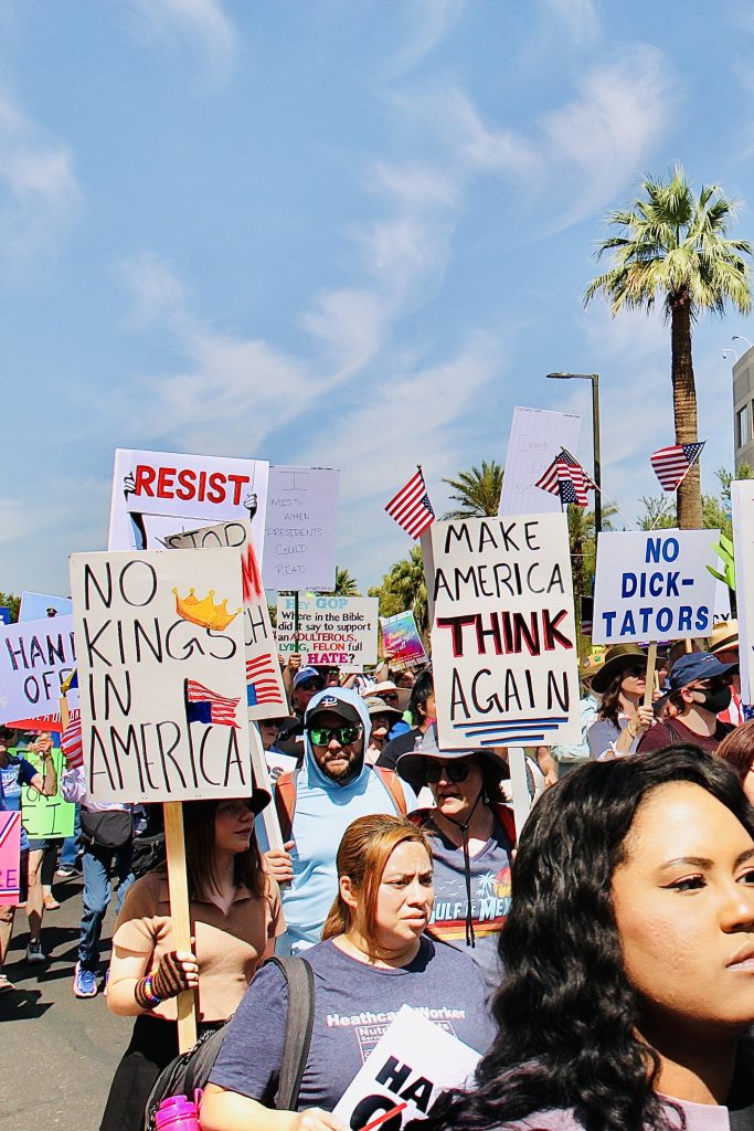 Protest march with about a dozen signs visible. Three completely readable: "No kings in America" "Make America THINK again" "No dick-tators" Three small U.S. flags visible.
