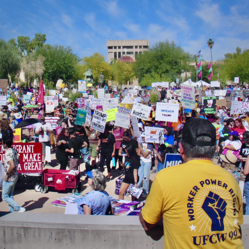 Dozens of protesters at the Arizona State Capitol holding up signs posing for a photo with a man in a yellow shirt looking at them.