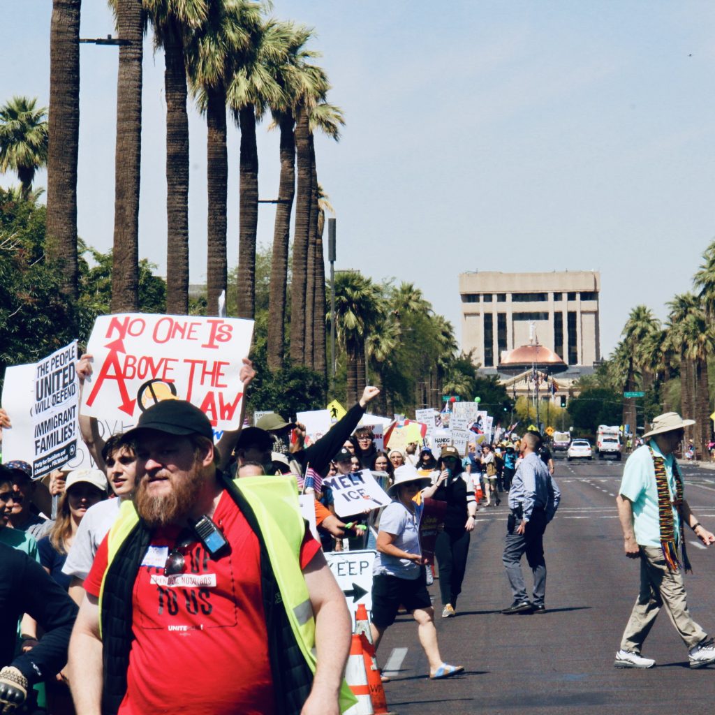 Wide view of protest march towards downtown Phoenix on the South side of Washington street. Arizona Capitol building visible in background. Protestors are holding signs.