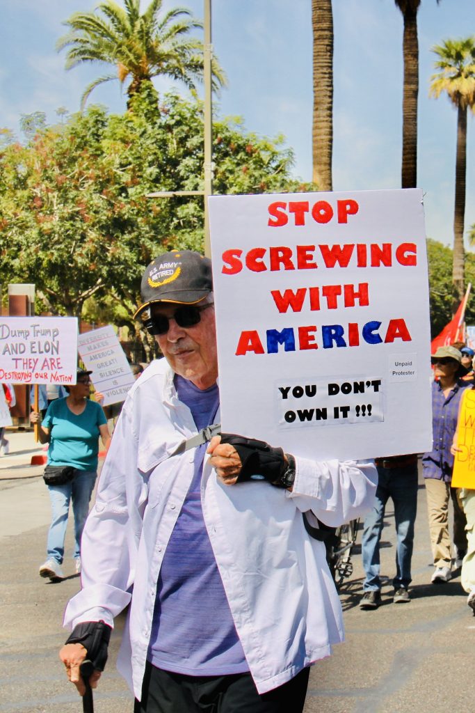 Protest march with a focus on an older man with "U.S> Army Retired" baseball cap and cane. Holding sign that says "Stop screwing with America" "You don't own it !!!" with smaller "Unpaid Protester" text.