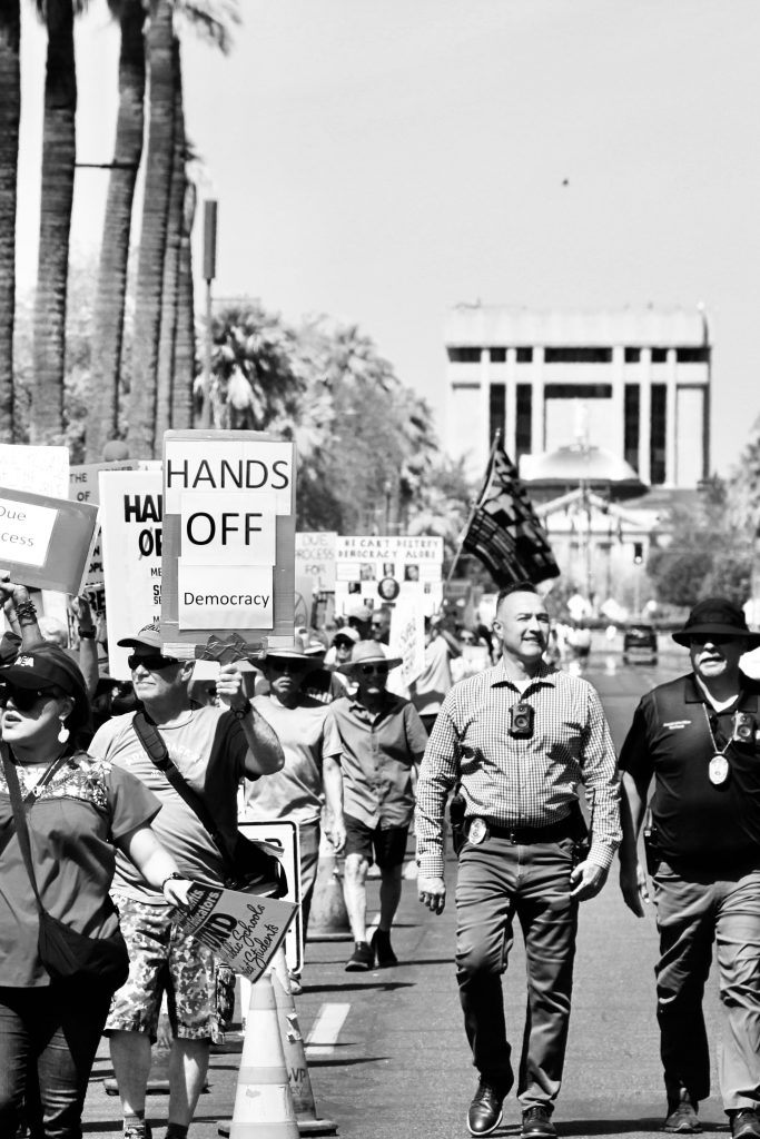 Black and white picture of protest march on the south side of Washington Street. Arizona Capitol Building visible in background. Two police officers with body cameras are also visible.