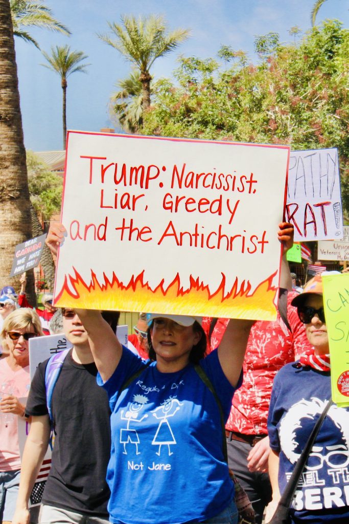 Protest march with a focus on a woman with a blue shirt with white stick figures on it. She is holding a sign that says "Trump: Narcissist, Liar, Greedy, and the Antichrist". White background, thin red outline, and flames on the bottom. Some sky and trees visible in the background.