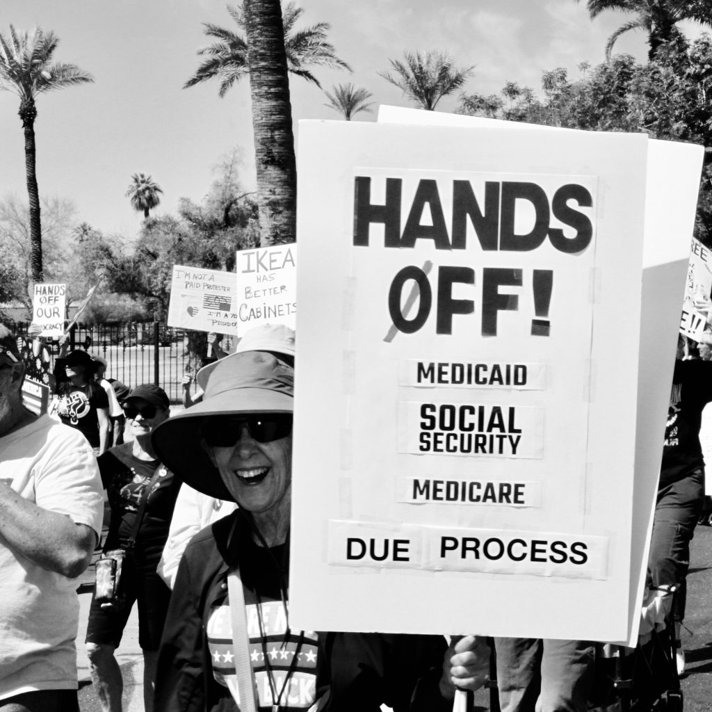 Black and white picture of protest march with focus on older woman in sun hat. She is holding a sign that says "Hands off!" (with cross through O) "Medicaid" "Social security" "Medicare" "Due Process"