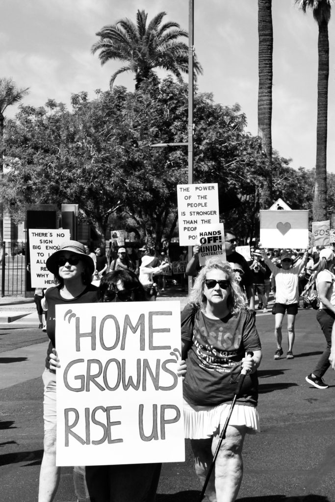 Black and white picture of protest march. Focus on two women holding sign that says "'HOME GROWNS' RISE UP"