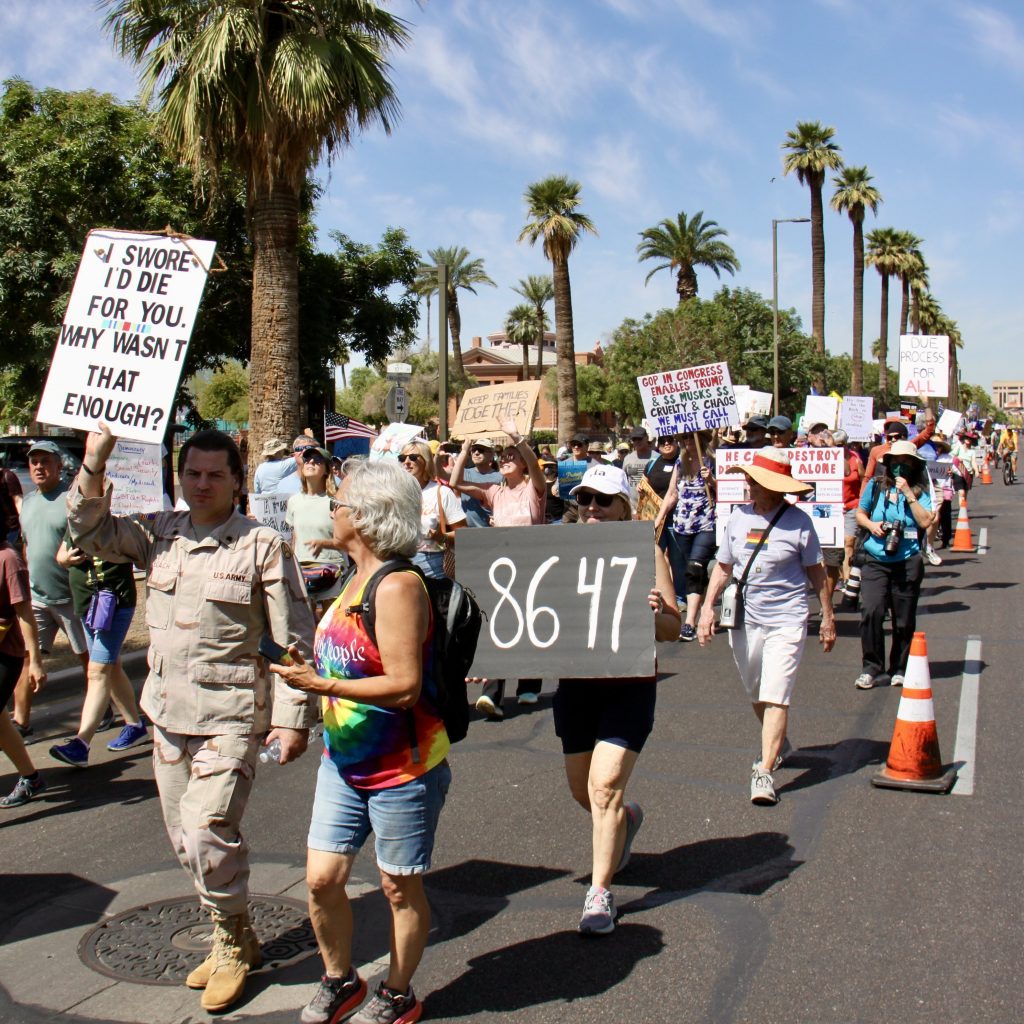 Protest march on South side of Washington Street towards Downtown Phoenix. Arizona State Capitol Building distant in background. Orange cones separating one lane. About a dozen signs visible.