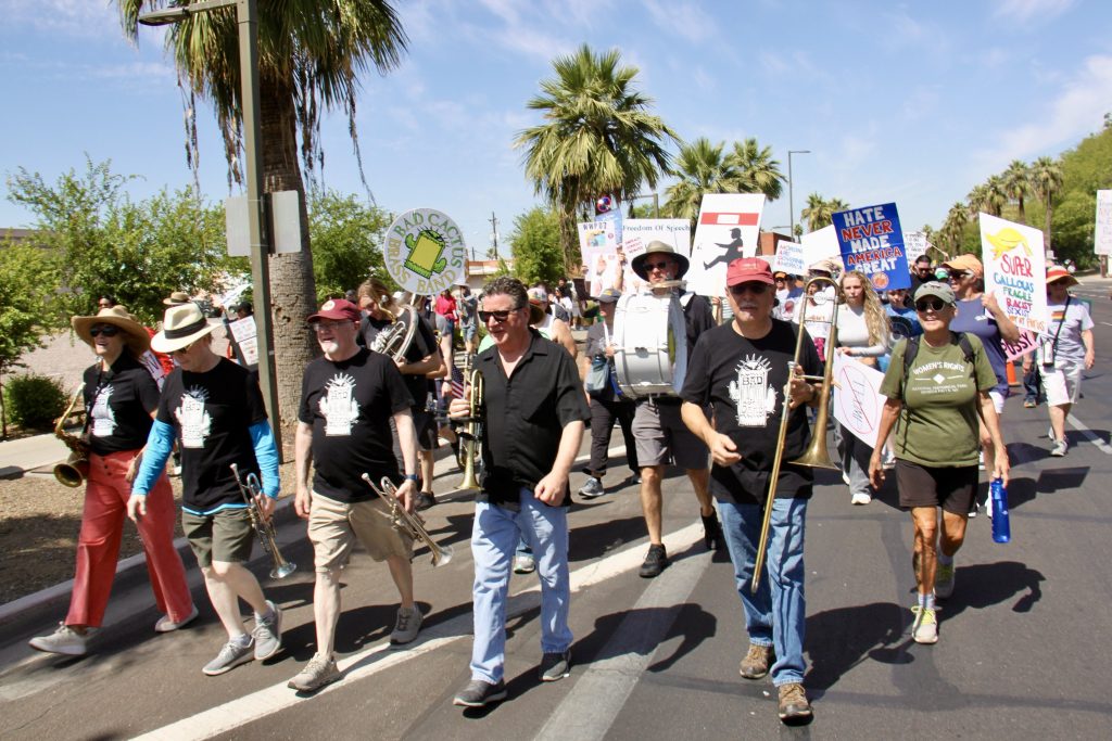 Protest march with five people carrying instruments in the front and a man with a drum mounted on his chest. About a dozen signs visible behind them.