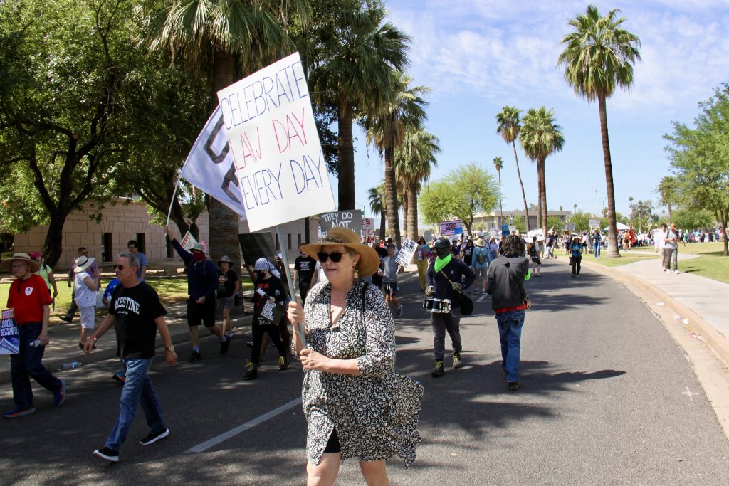 Picture of protest march heading East from the Arizona State Capitol, focusing on a woman holding a sign that says "CELEBRATE LAW DAY EVERY DAY"