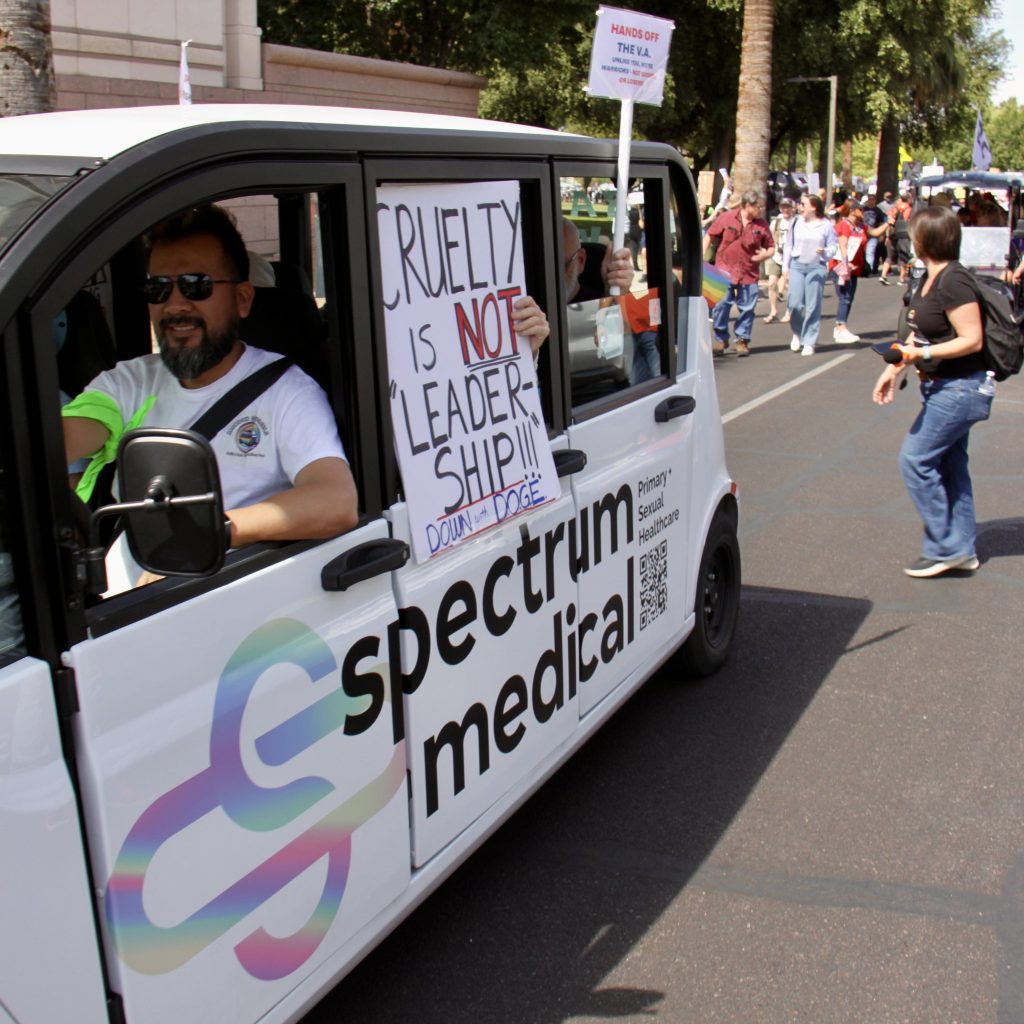A small vehicle driving alongside a protest march with "spectrum medical" logo on it. Viewed from front left of vehicle. Driver is smiling and wearing sunglasses. Protestors are holding signs outside the vheicle: "Cruelty is _NOT_ 'leader-ship!!!' DOWN with DOGE" and "Hands off the V.A. Unlike you we're warriors, not suckers and loosers."